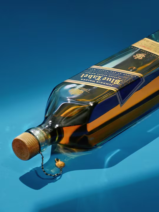 Una botella de whisky Johnnie Walker Blue Label está tumbada sobre un fondo azul brillante. La botella está cerrada con un tapón dorado y lleva un emblema decorativo en la etiqueta.