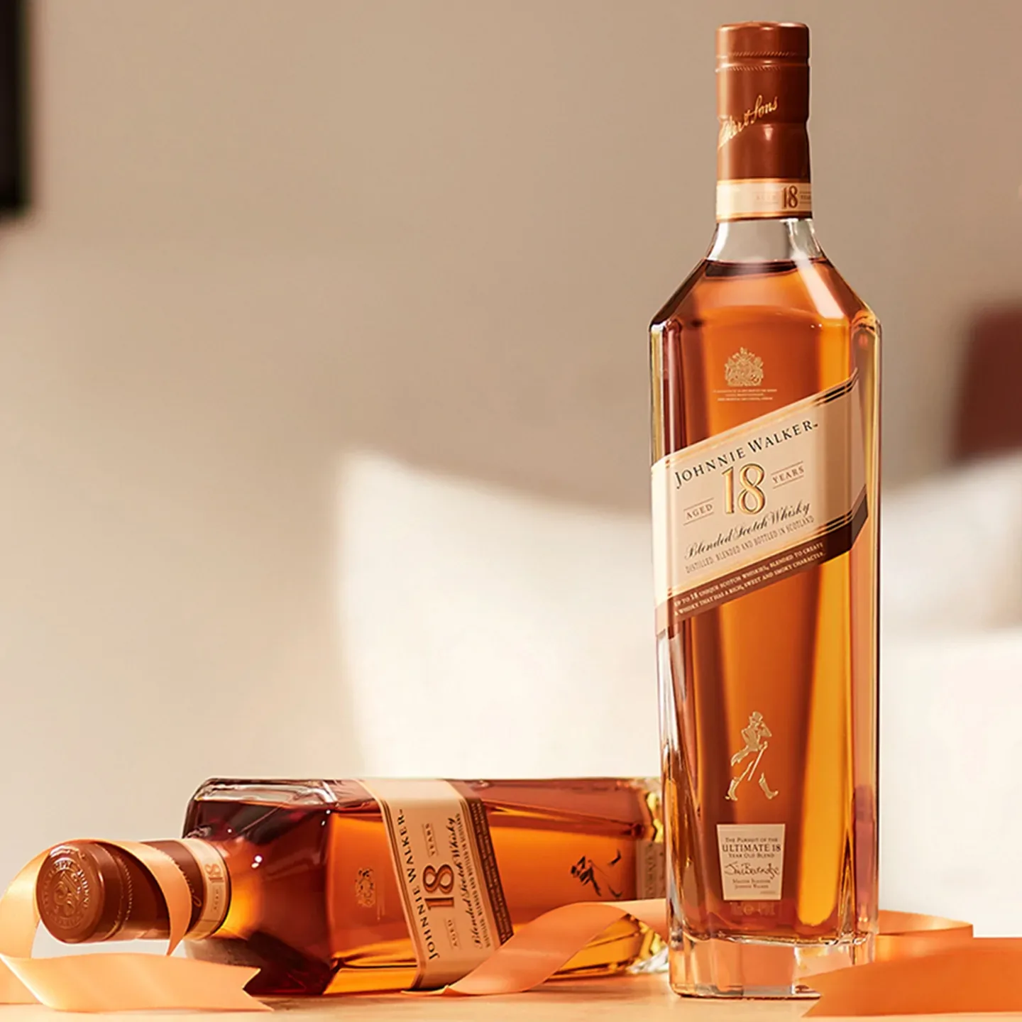 Two bottles of Johnnie Walker 18-Year-Old Scotch Whisky are displayed. One bottle is upright, and the other is lying on its side with a ribbon draped around it, against a blurred indoor background.