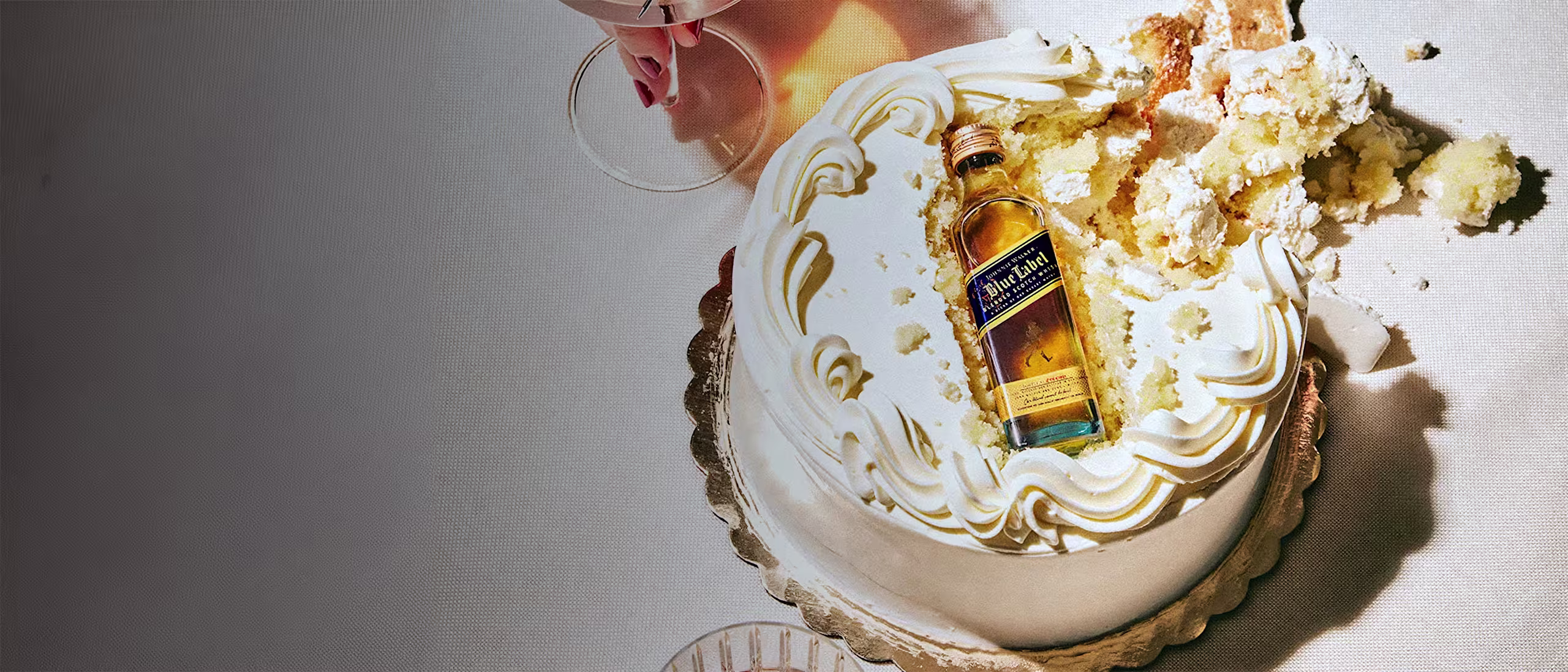 A small cake with white frosting is partly smashed, revealing a miniature bottle of whisky inside. Next to the cake are a red cocktail with berries and another glass with an amber drink.