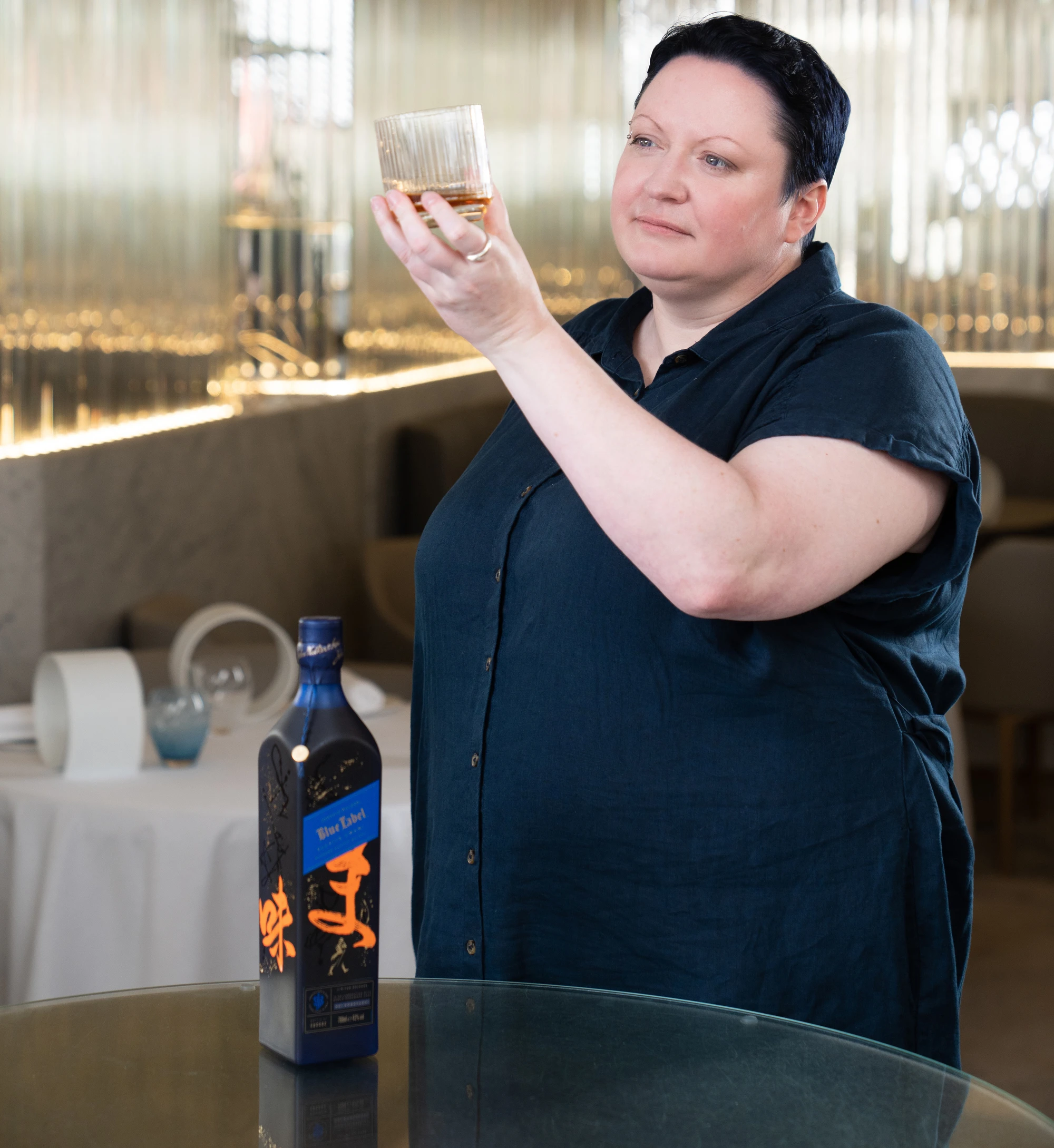 A woman in a blue shirt holds up a glass of whiskey and a bottle of Blue Label whiskey sits on a table in front of her. 
