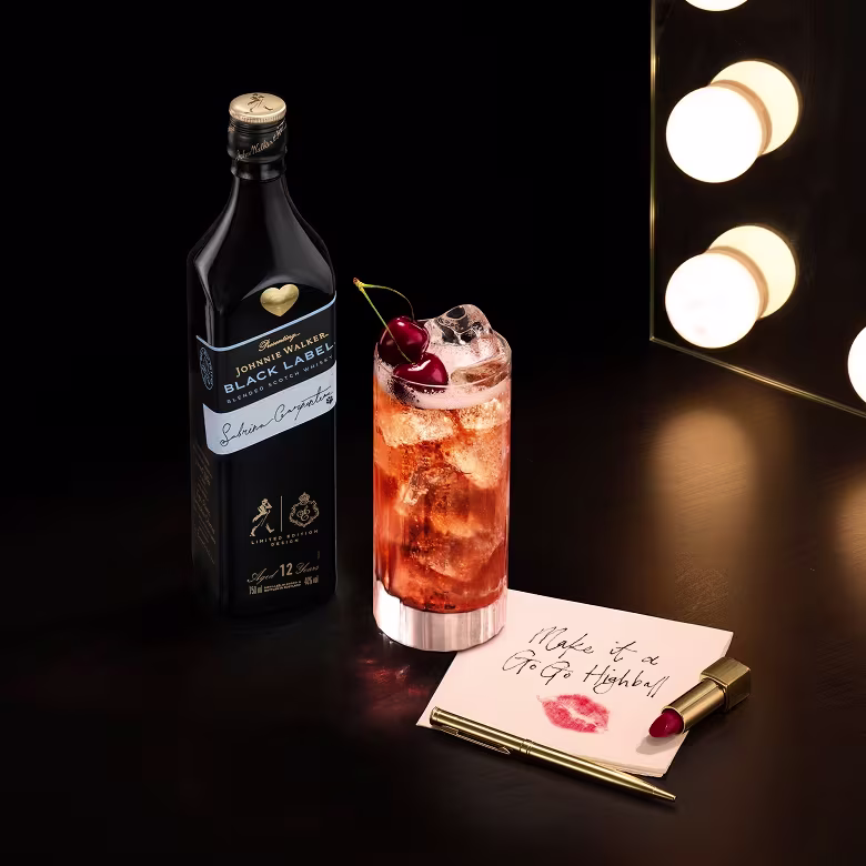 Bottle of Johnnie Walker Black Label, a cocktail with a cherry, lipstick, and a note on a dark vanity table.