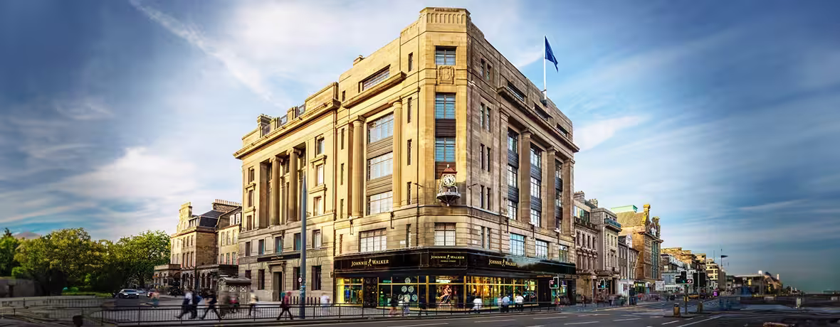 A grand, multi-story stone building with art deco architecture stands on a street corner under a blue sky. A blue flag flies from the roof, and shops with large windows line the ground floor.