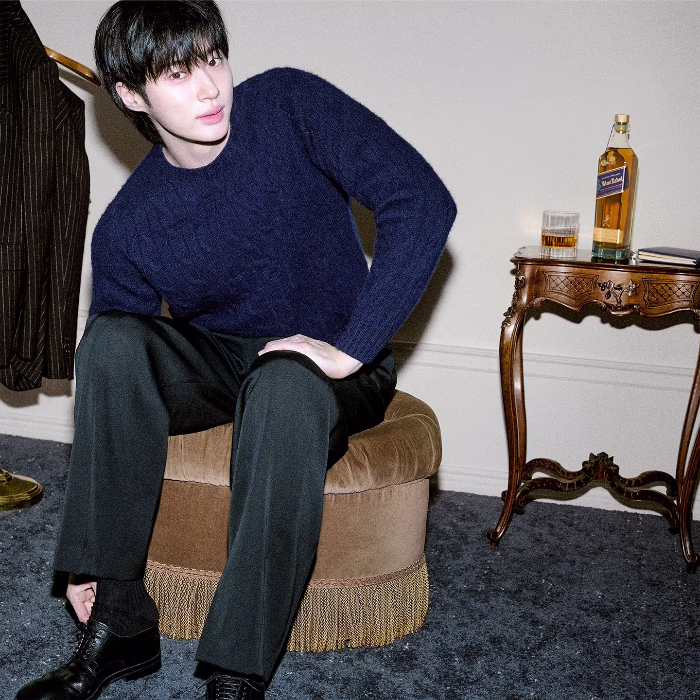 A person in a navy sweater sits on a round chair, tying a shoe, with a whiskey bottle on a side table nearby.
