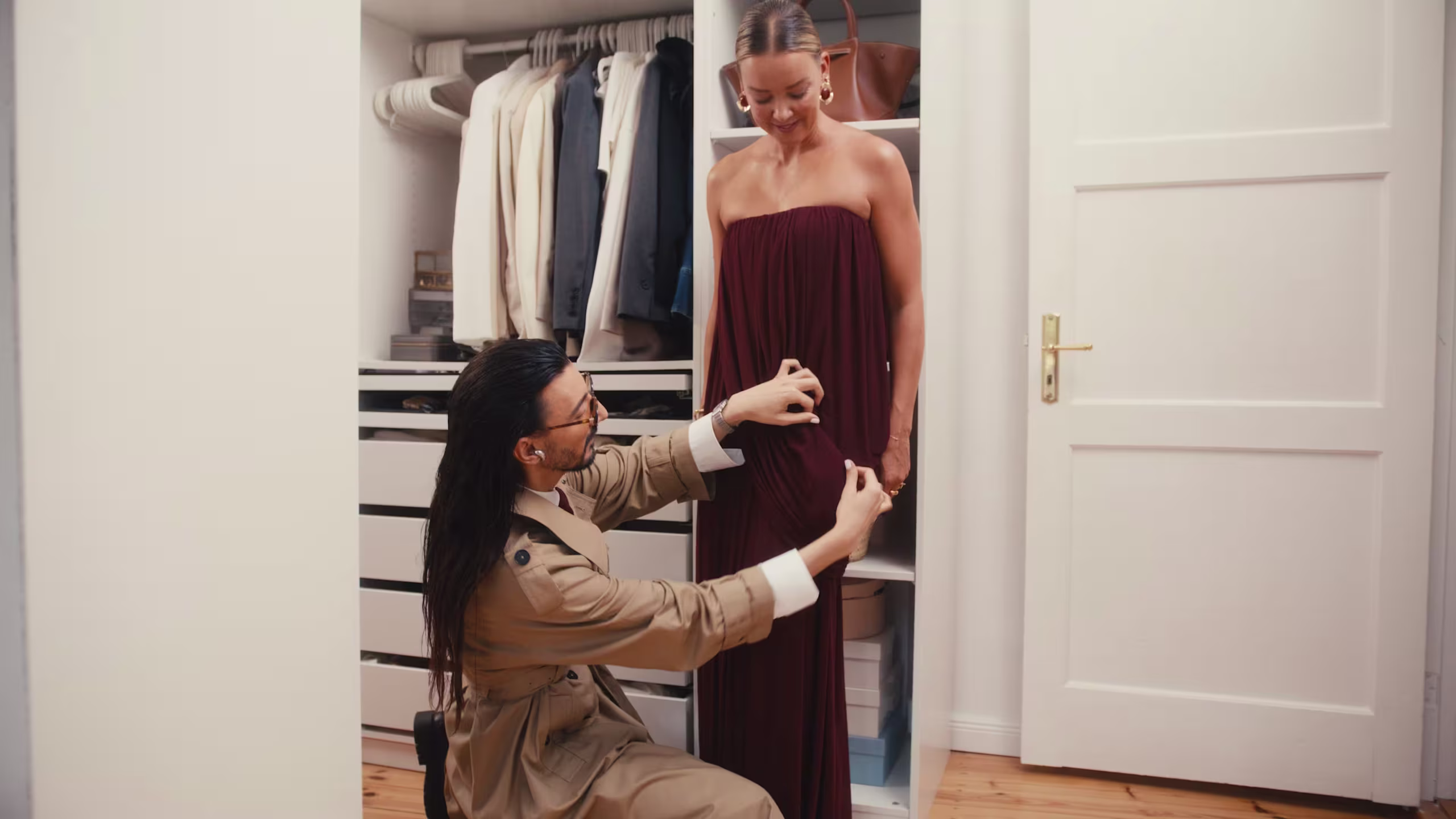 Eine Modedesignerin kniet nieder, um einer lächelnden Frau, die in einem begehbaren Kleiderschrank voller hängender Kleidung und Regalen steht, ein langes, trägerloses kastanienbraunes Kleid anzupassen.