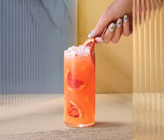 A hand with colorful nail art holds a tall glass of orange cocktail topped with ice and garnished with citrus slices. The setting includes textured glass panels and a neutral-toned surface.