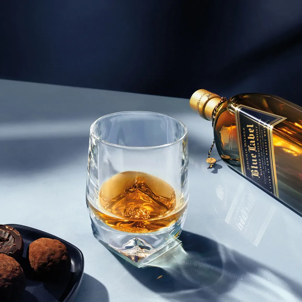 A glass filled with whiskey sits on a reflective surface next to an open blue label whiskey bottle. A plate with assorted dark chocolates is nearby, creating a sophisticated scene under soft lighting.