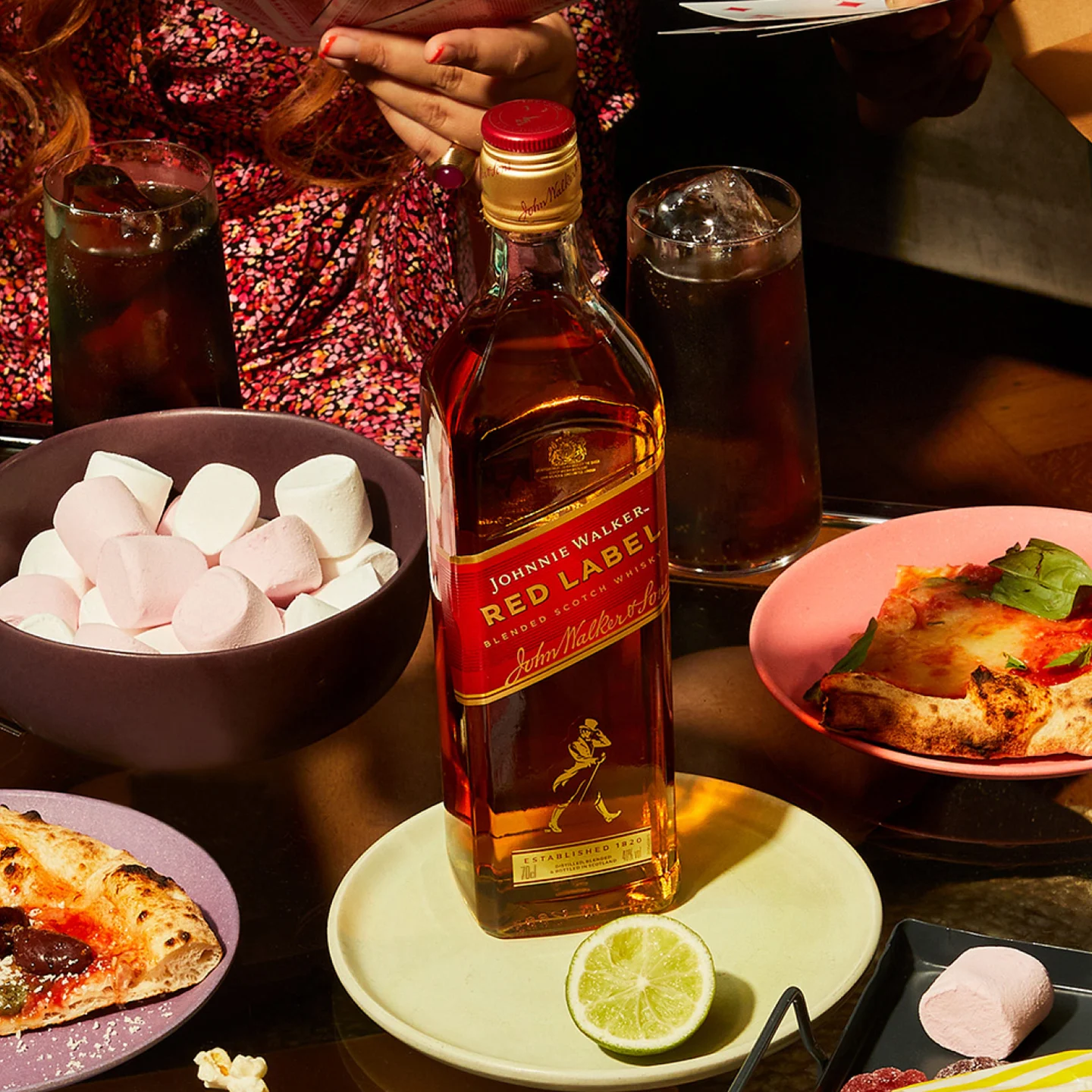 Ένα μπουκάλι ουίσκι Johnnie Walker Red Label σε ένα τραπέζι που περιβάλλεται από μια φέτα λάιμ, φέτες πίτσας, μπολ με marshmallows και δύο ποτήρια γεμάτα με σκούρο υγρό. Ένα άτομο είναι μερικώς ορατό κρατώντας κάρτες στο παρασκήνιο.