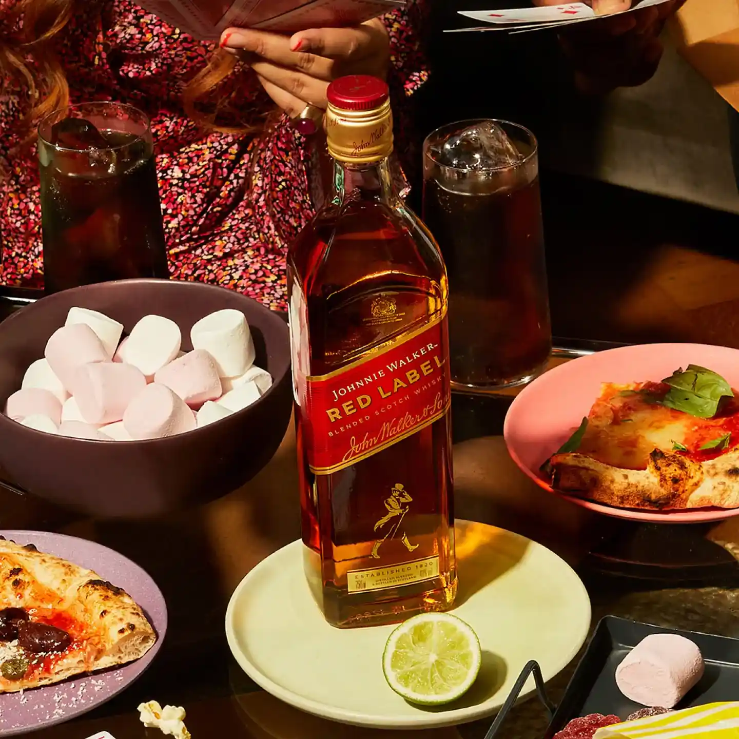 Een fles Johnnie Walker Red Label whisky staat op een tafel, omringd door schalen vol marshmallows, stukken pizza, glazen frisdrank, een schijfje limoen en een hand die speelkaarten vasthoudt.