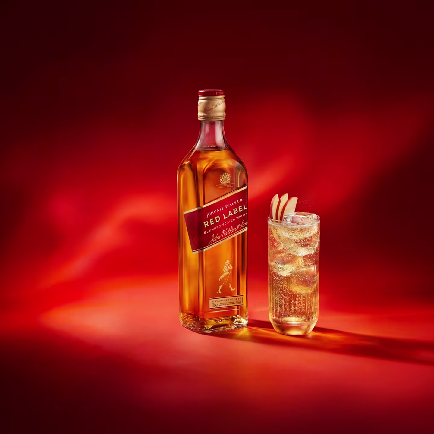 Una botella de whisky Johnnie Walker Red Label junto a un vaso de whisky con hielo y una rodaja de limón sobre un fondo rojo.