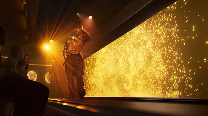  Un actor con sombrero de copa y traje teatral se encuentra en un escenario iluminado por luces doradas y efectos brillantes, mientras el público observa desde un lado.