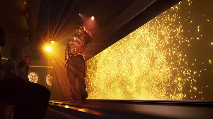  Un artiste portant un chapeau haut de forme et un costume de théâtre se tient sur une scène éclairée par des lumières dorées et des effets scintillants, tandis qu'un public regarde de côté.