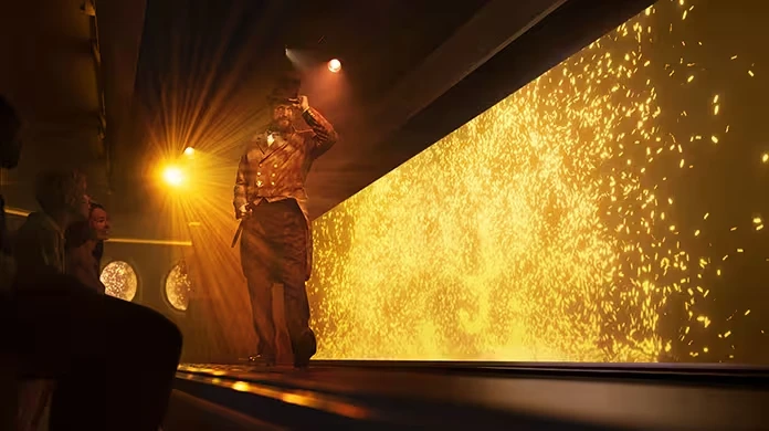 A performer in a top hat and theatrical costume stands on a stage lit by golden lights and sparkling effects, while an audience watches from the side.