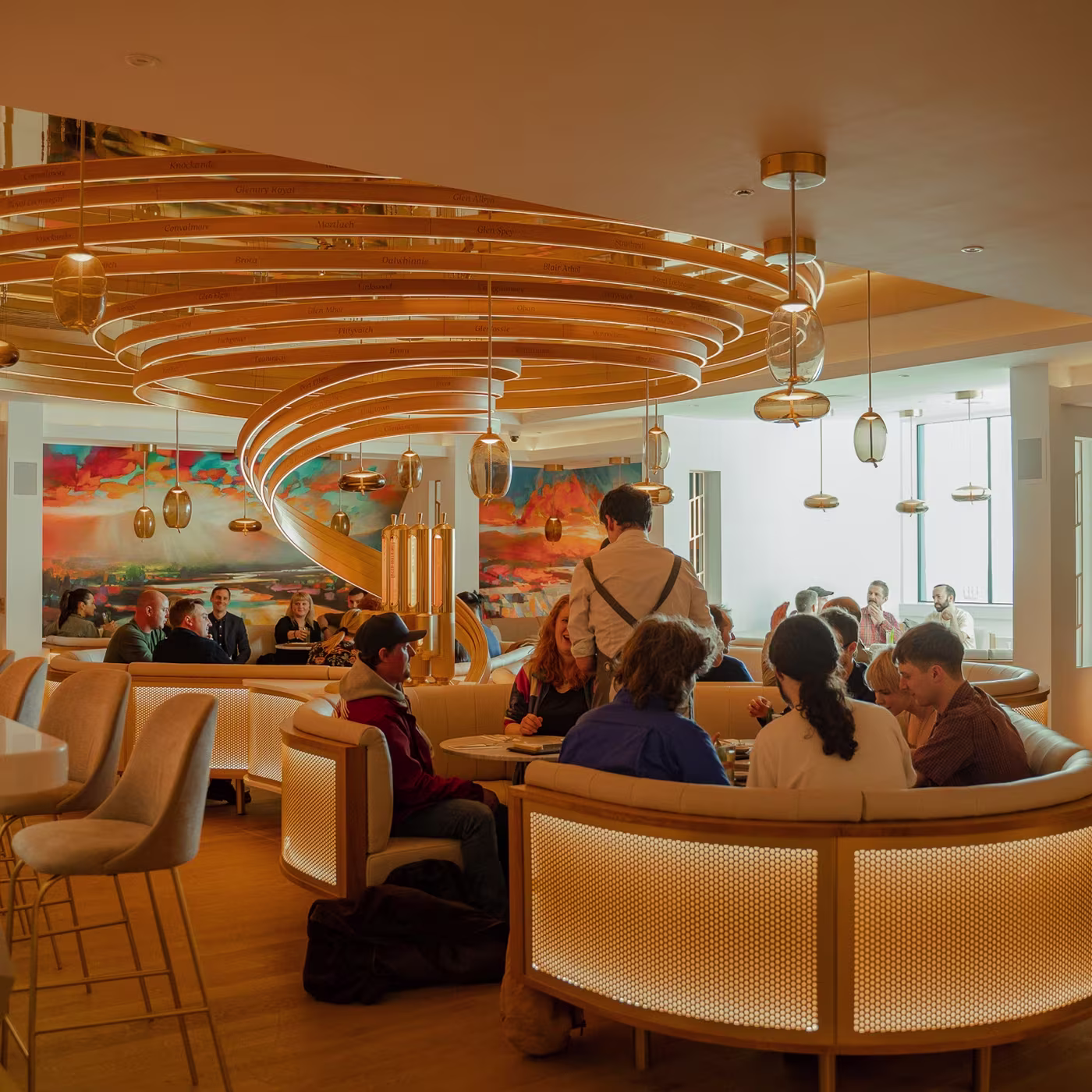 A modern restaurant with a spiral staircase, golden lighting, and people dining in cozy booths.