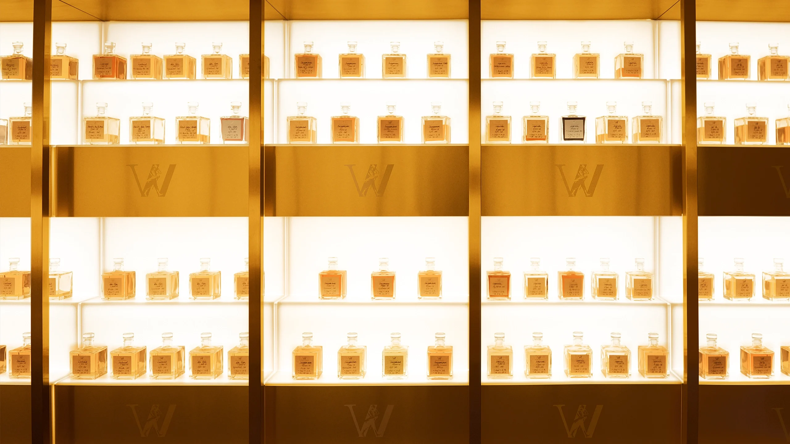 A well-lit display of perfume bottles arranged in neat rows and columns on a golden shelf. Each shelf has a warm glow, highlighting the individual bottles. The backdrop features a metallic letter W at intervals.
