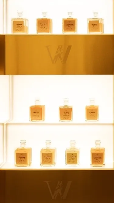 A well-lit display of perfume bottles arranged in neat rows and columns on a golden shelf. Each shelf has a warm glow, highlighting the individual bottles. The backdrop features a metallic letter W at intervals.