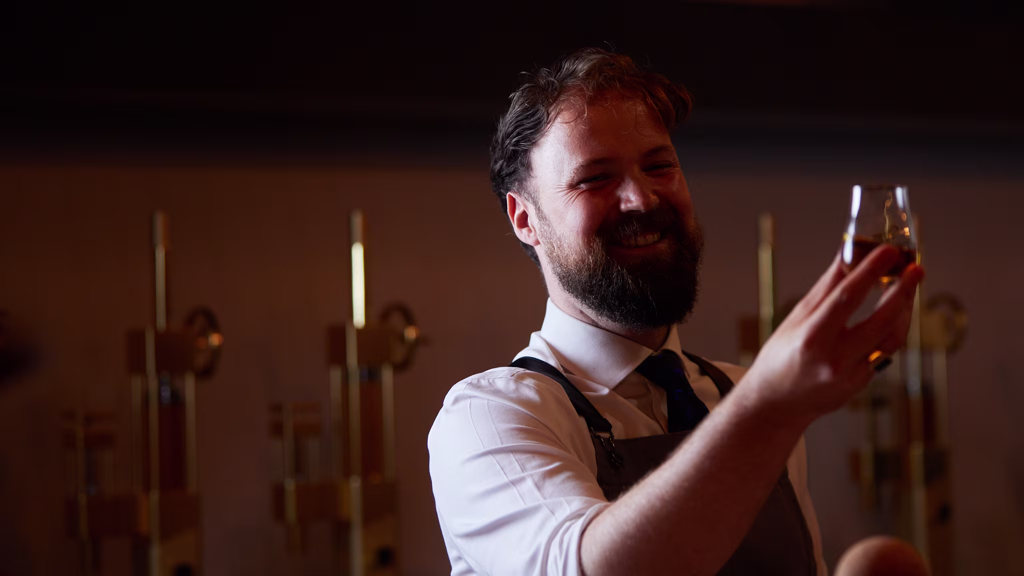 A bearded man in a white shirt and apron smiles while holding up a glass of whiskey for inspection in a dimly lit room with golden fixtures.