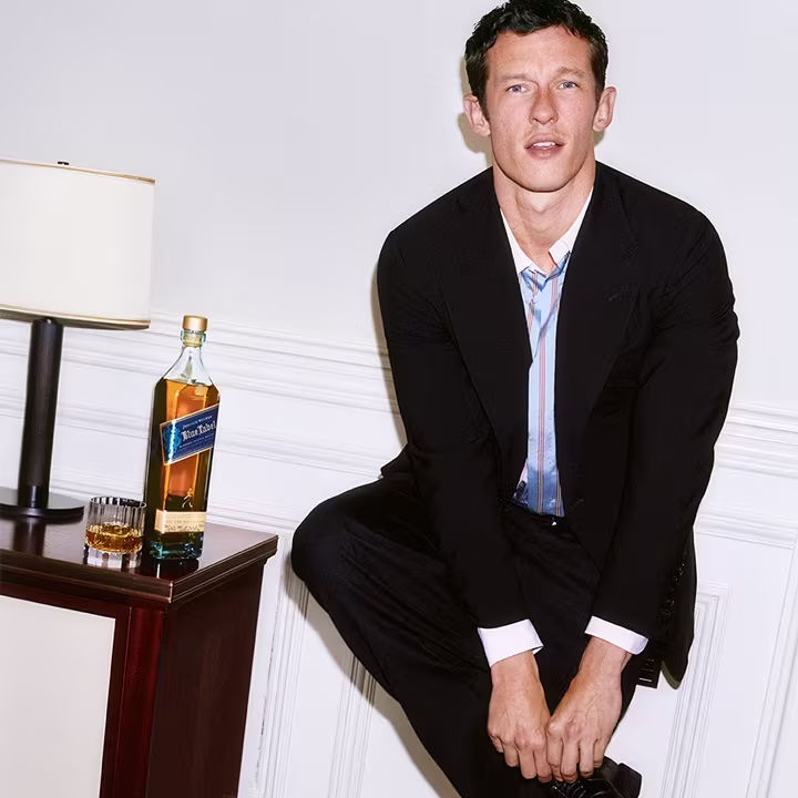 Callum Turner in suit sitting beside a table with a bottle of whisky, glass, and lamp in a bright room.