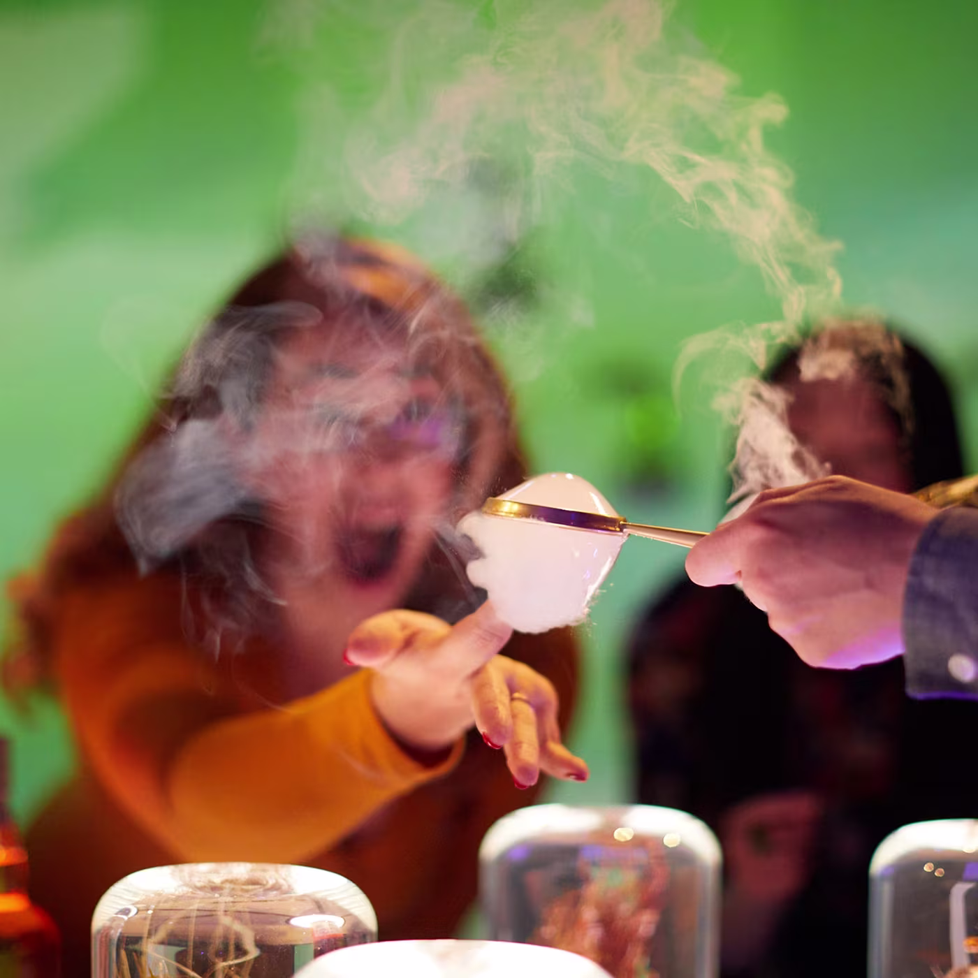A woman reaches excitedly toward a smoky bubble held by tongs against a green background.