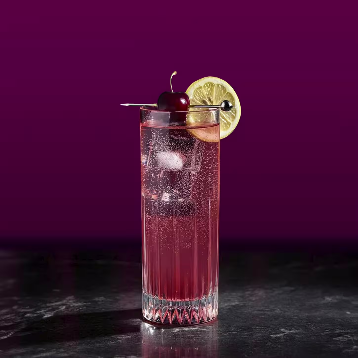 Un vaso alto de una bebida gaseosa roja con hielo está adornado con una rodaja de limón y una cereza en un palillo de cóctel, sobre un fondo violeta oscuro sobre una superficie de mármol negro.