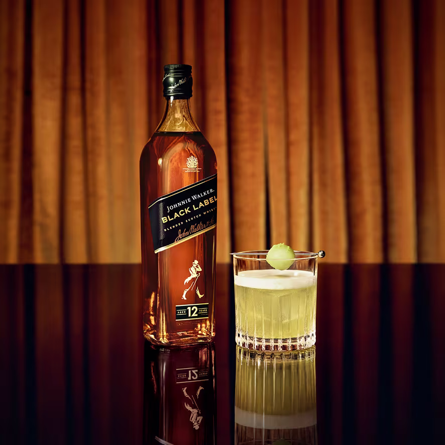 琥珀色布幕前，岩石杯中放著哈密瓜球的 TOXIC SOUR 調酒，旁邊放著一瓶 Johnnie Walker 黑牌。