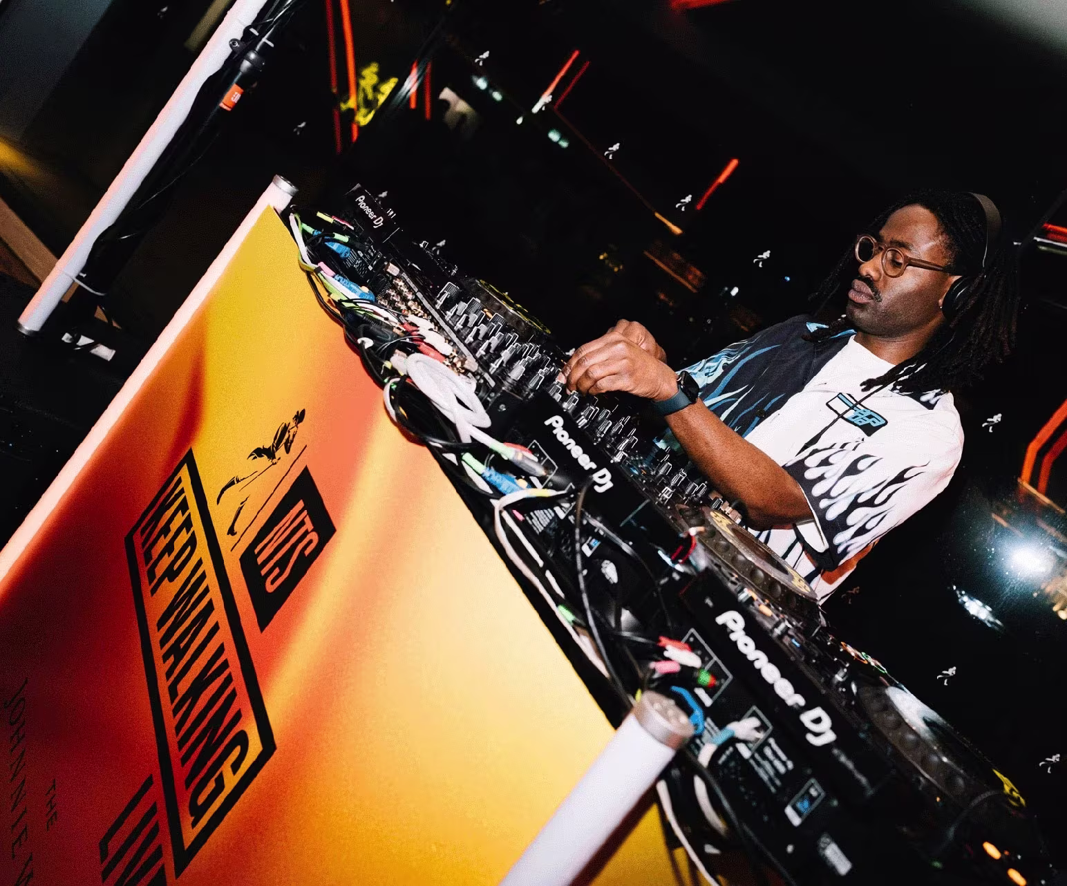 A DJ wearing glasses performs at a brightly lit booth with audio equipment and a yellow sign.