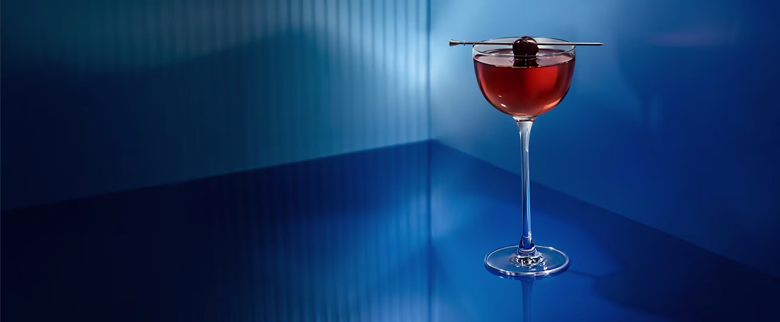 Blue Manhattan cocktail glass with a dark drink and cherry garnish sits on a shiny blue surface with dramatic lighting.
