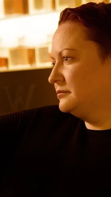 Person with short hair holds a glass container with amber liquid, examining it intently. Shelves filled with bottles are in the background, illuminated with warm lighting.