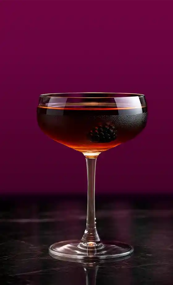 A coupe glass filled with a dark liquid and a blackberry garnish, set against a rich purple background. The glass sits on a dark surface, creating a sophisticated and elegant presentation.