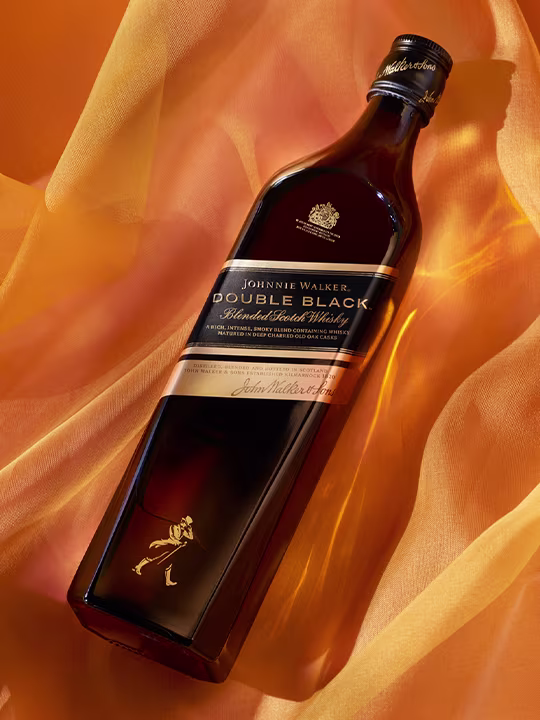 Бутилка смесено шотландско уиски Johnnie Walker Double Black с тъмен етикет със златен текст и лого. Бутилката е поставена на фона на течащ златен плат.