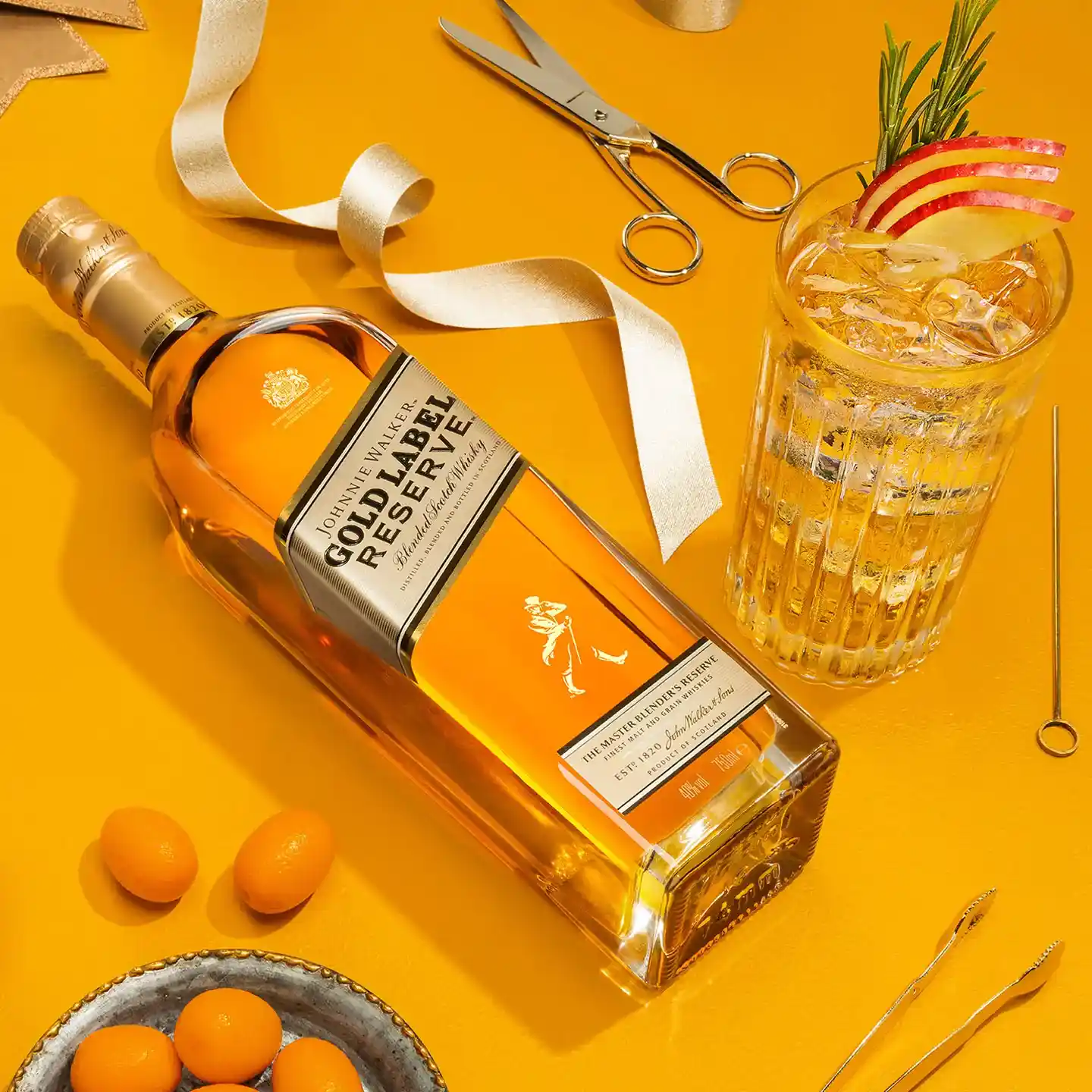 A bottle of Gold Label Reserve whiskey lies on a yellow surface next to a glass filled with a drink garnished with apple slices and rosemary. Surrounding items include ribbon, scissors, a dish of kumquats, and a cocktail stirrer.