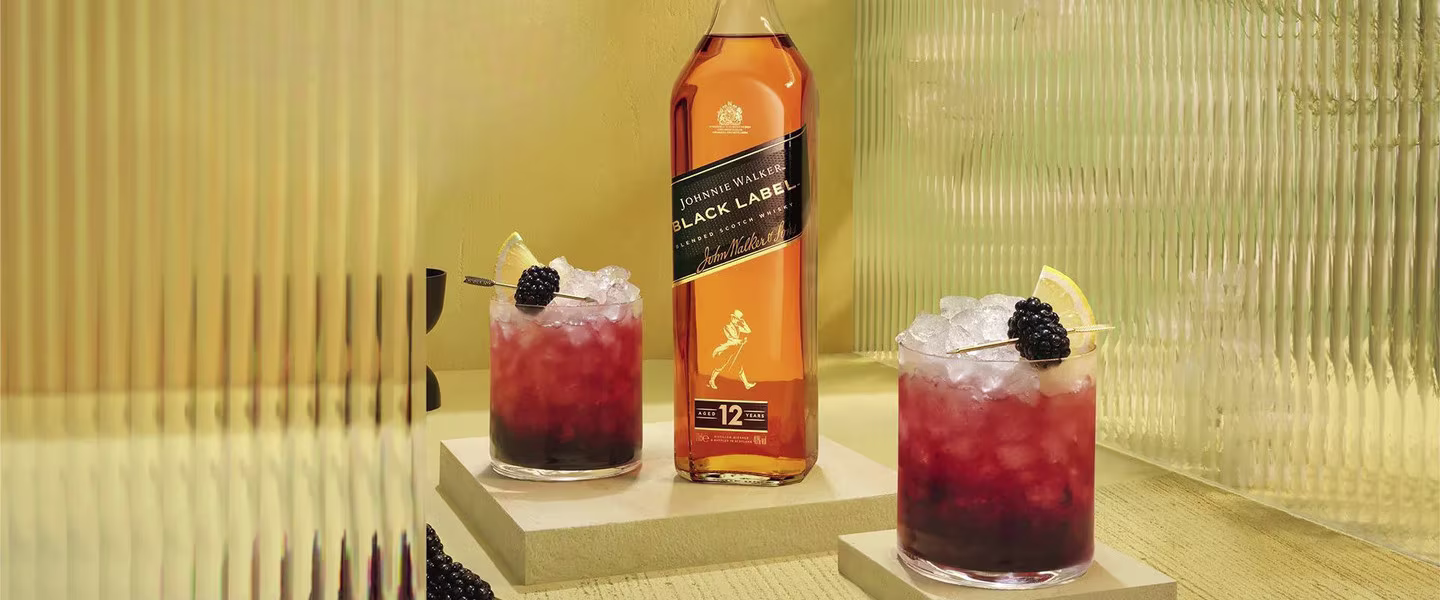 A bottle of Johnnie Walker Black Label sits between two whiskey cocktails on stone blocks. Each drink is garnished with a lemon wedge and a blackberry on a skewer. The background is a textured, golden surface.