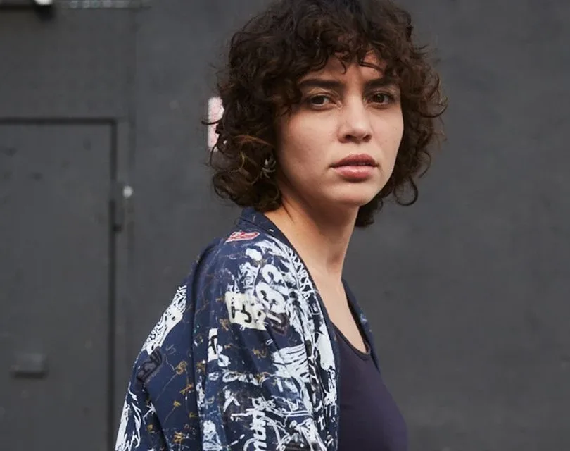 Uma pessoa com cabelos cacheados, vestindo uma camisa estampada, está em frente a um fundo cinza escuro, olhando para a câmera com uma expressão neutra.