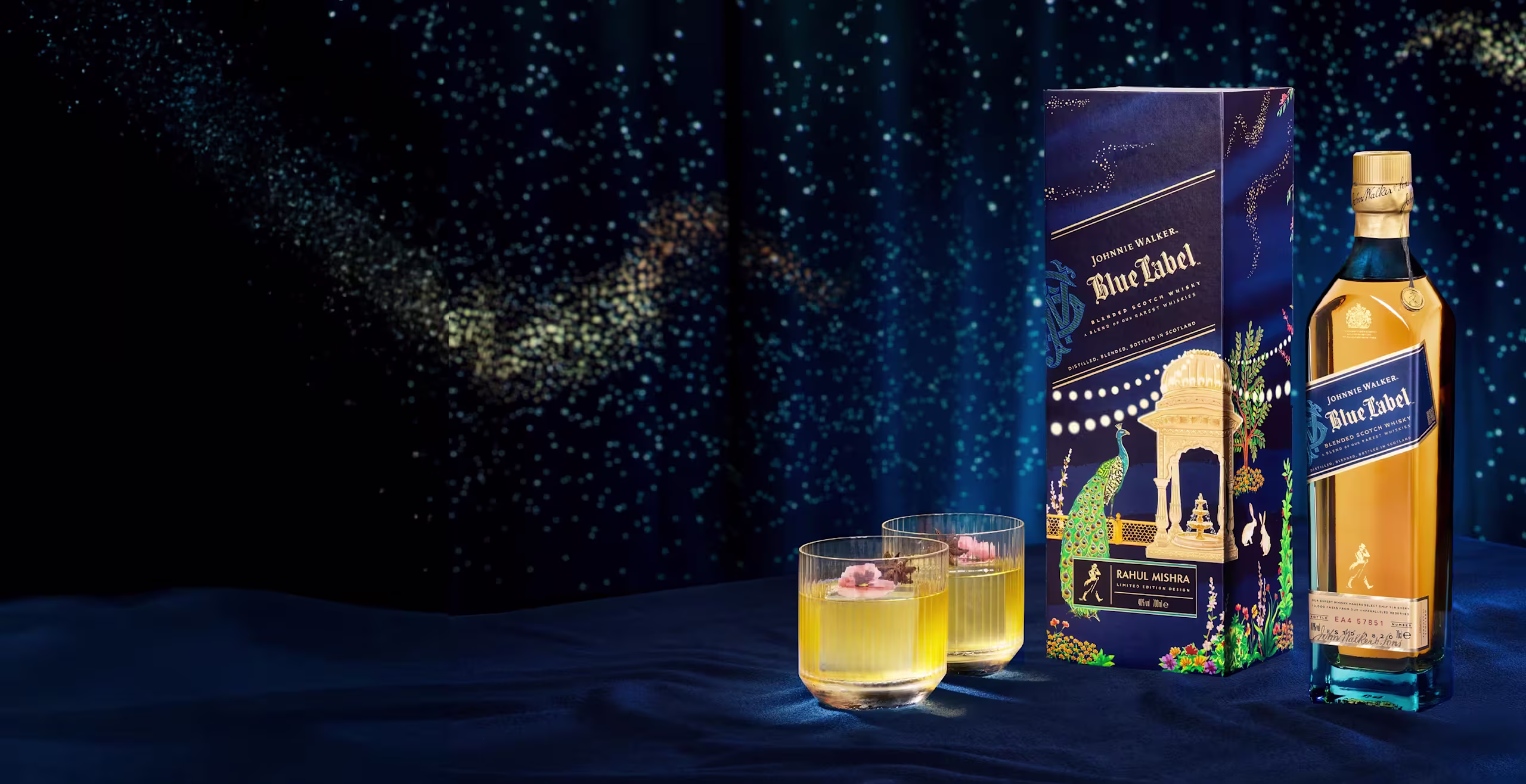 A bottle of Johnnie Walker Blue Label with a decorative box and two yellow cocktails garnished with pink flowers, set on a blue surface with a starry, dark blue background.