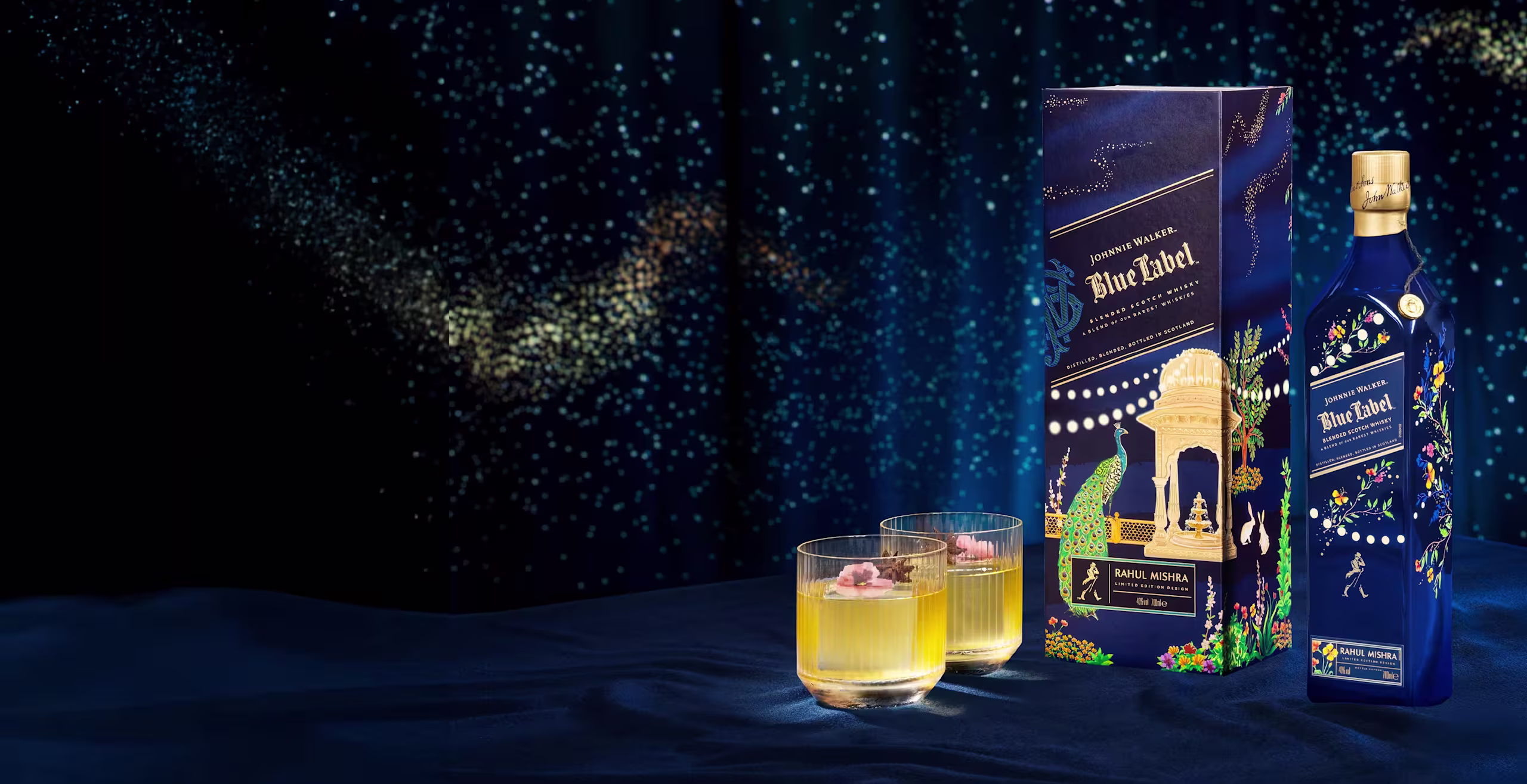 A bottle of Johnnie Walker Blue Label X Rahul Mishra Limited Edition whiskey with a decorative blue box and two whiskey glasses with ice and garnishes, set on a dark blue fabric with a starry background.