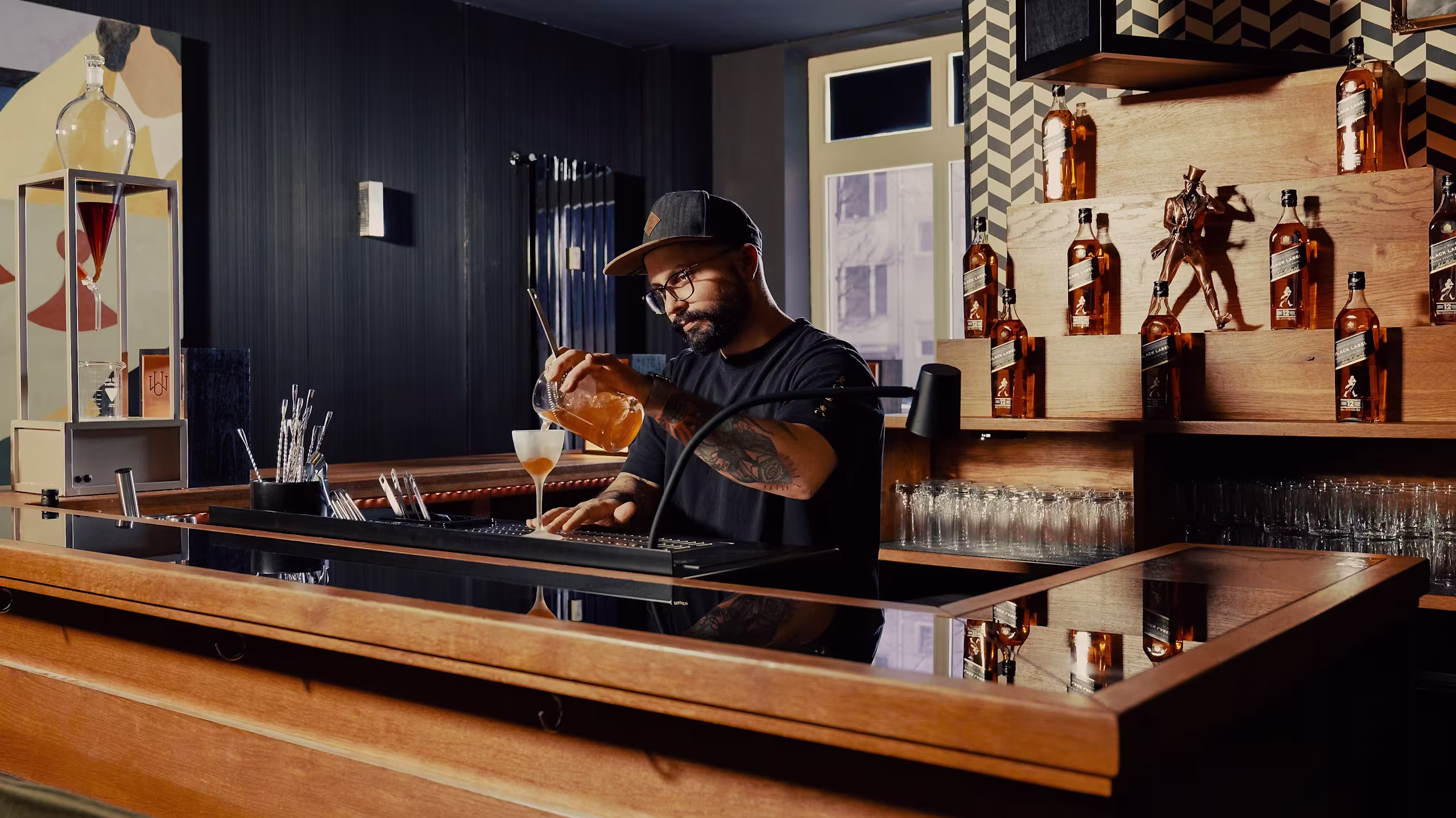 Ein Barkeeper mit Brille und Mütze schenkt hinter einer modernen Bar einen Drink ein, umgeben von Whiskyflaschen und Cocktailgläsern, im Hintergrund eine geometrische Wanddekoration.