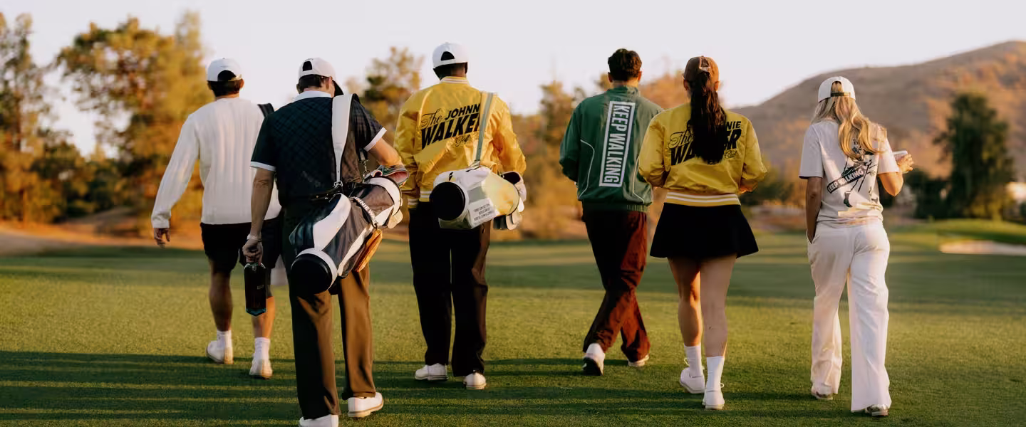 Five people in casual sportswear are walking on a golf course with clubs. They wear yellow and green jackets with text, and it's sunny with a clear sky. Trees and hills in the background.