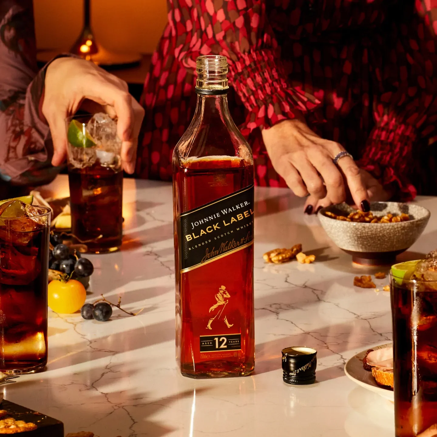 Una botella de whisky Johnnie Walker Black Label reposa sobre una mesa de mármol rodeada de vasos llenos de bebidas. Dos manos se estiran para coger aperitivos y guarniciones. La escena está iluminada de forma cálida, lo que crea una atmósfera acogedora.