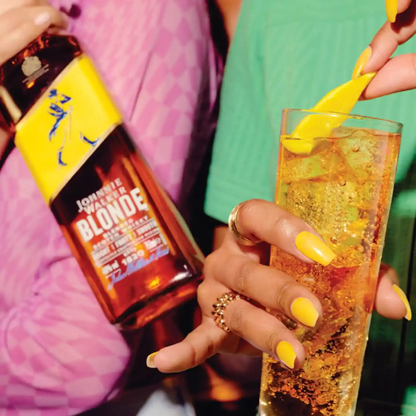 Una persona con las uñas pintadas de amarillo sostiene un cóctel con una rodaja de limón como decoración, mientras que en otra mano sostiene una botella de Johnnie Walker Blonde. 