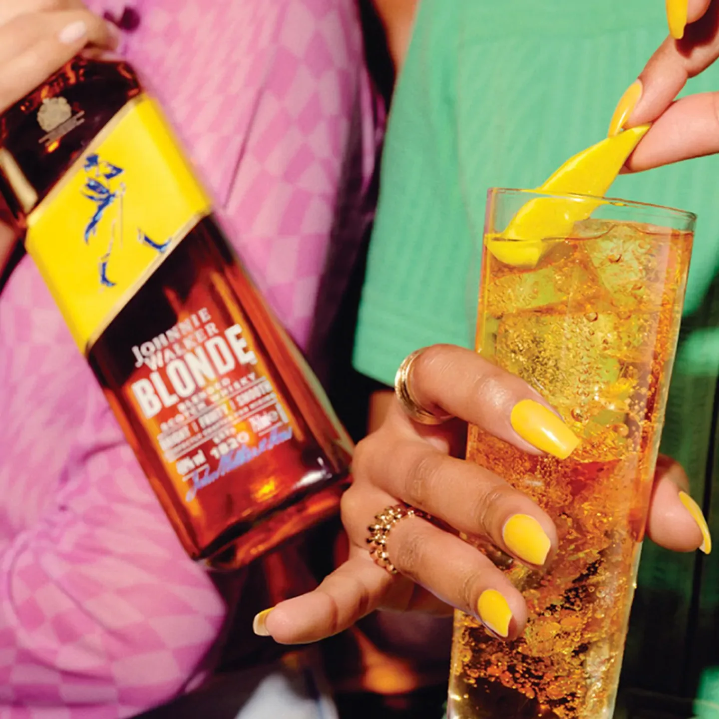 A person with yellow nail polish holds a glass filled with an amber-colored drink and ice, garnished with a yellow peel. Another hand holds a bottle labeled Johnnie Walker Blonde. The background is blurred with colorful clothing.