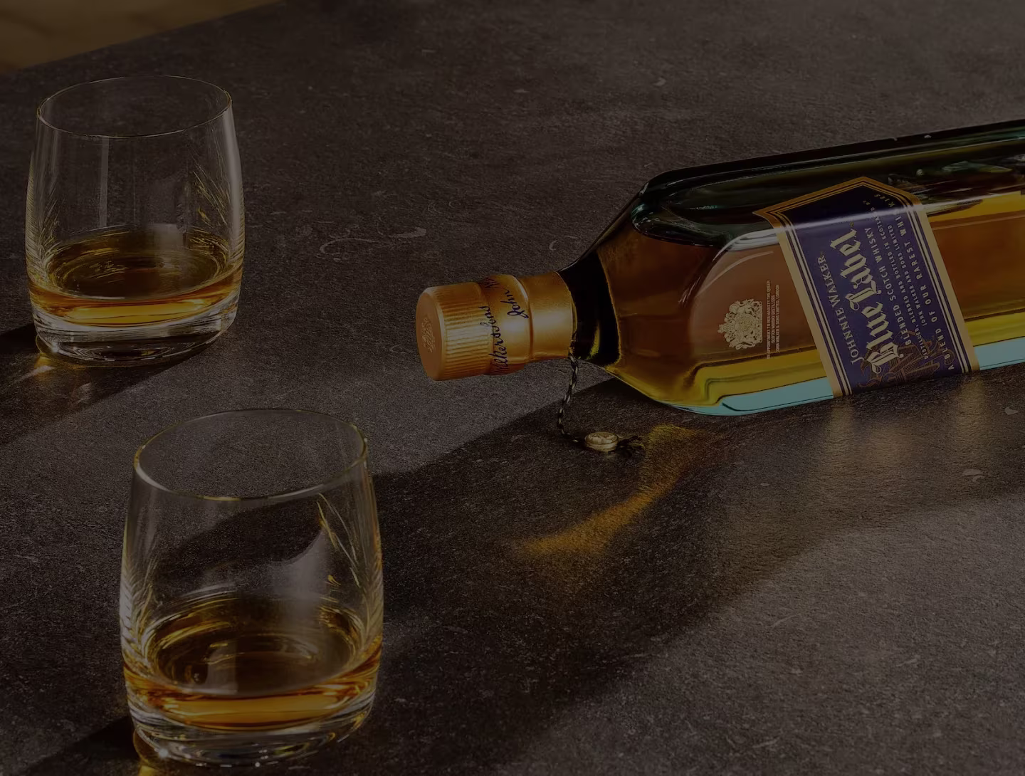 Une bouteille de whisky Blue Label Johnnie Walker repose sur le côté, coulant sur un comptoir sombre, avec deux verres de whisky à proximité.
