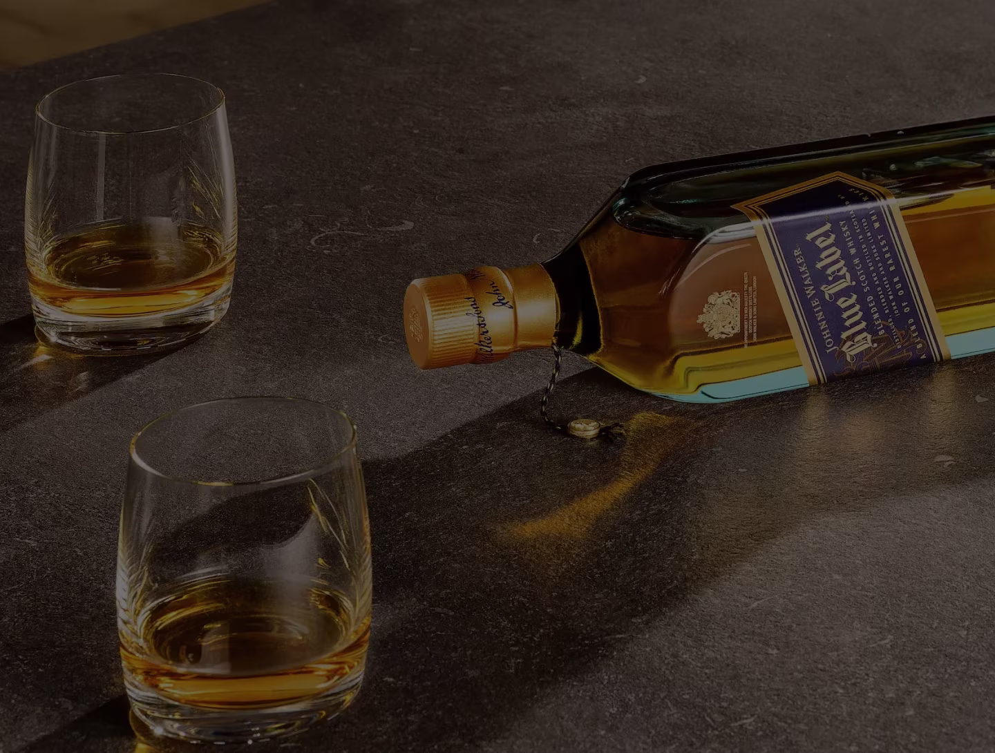 Una Johnnie Walker Blue Label botella de whisky se encuentra volcada y su contenido se derrama sobre una superficie oscura. Cerca hay dos vasos de vidrio, uno casi lleno y otro con algo de líquido, que reflejan cálidos tonos ámbar.