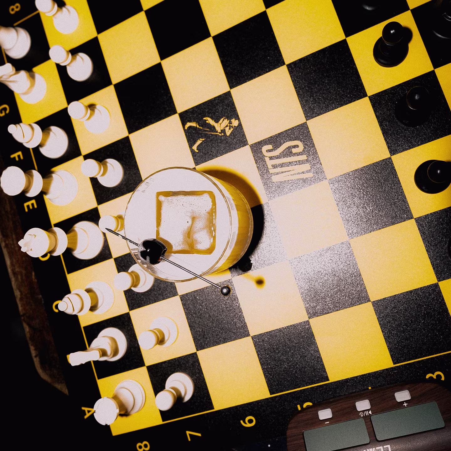 A cocktail with ice sits on a black and yellow chessboard surrounded by white and black chess pieces.