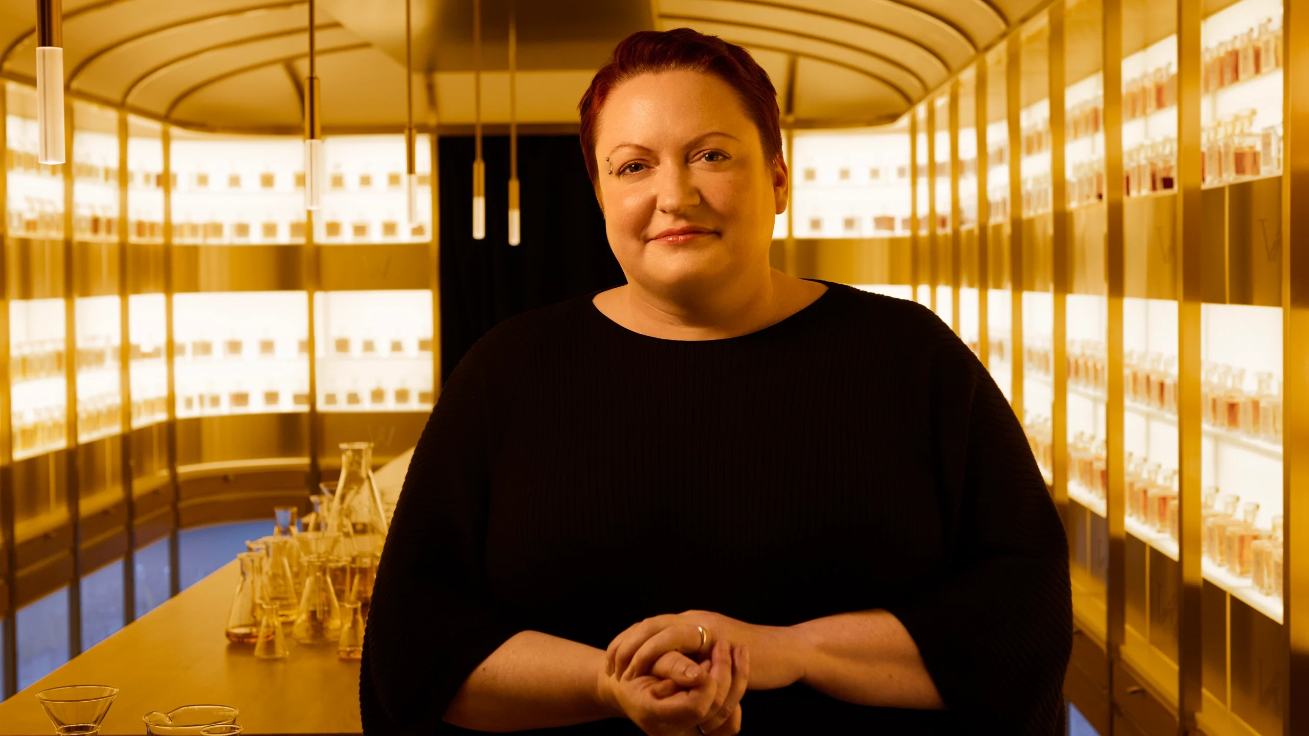 Dr. Emma Walker with short hair standing in a warmly lit room full of shelves lined with bottles. They are wearing a black top, with hands clasped. Tables with glassware are visible in the background.