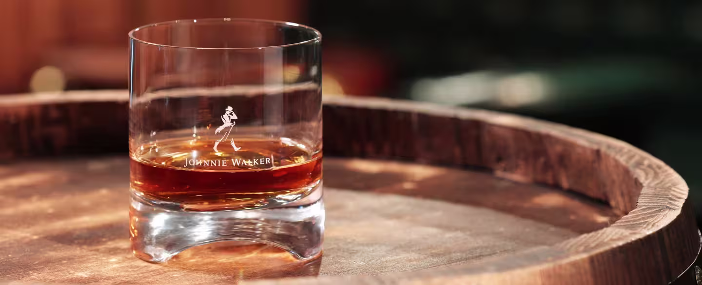 A glass of Johnnie Walker whisky, featuring the brand’s logo, sits on a wooden barrel in a warmly lit setting.