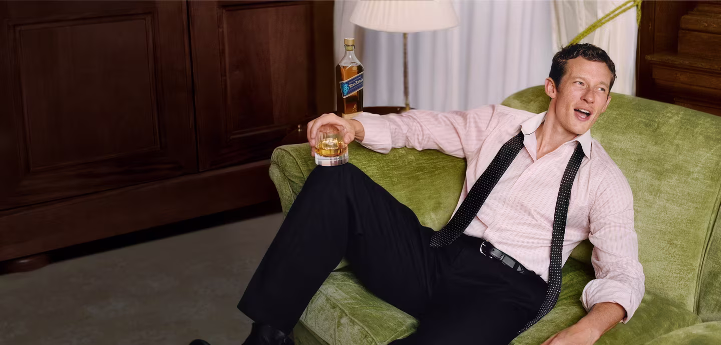 Callum Turner relaxing on a green couch, holding a glass of whiskey, with a whiskey bottle on the table behind him.
