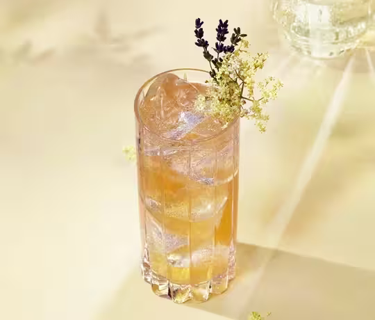 Un vaso alto lleno de una bebida de color ámbar claro, cubitos de hielo y adornado con pequeñas flores blancas y una ramita de lavanda, reposa sobre una superficie pálida bañada por una suave luz natural. Se ven sombras de flores en el fondo.