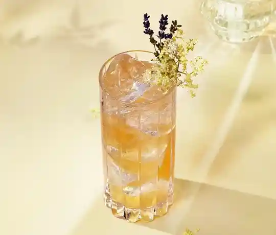 Un vaso alto lleno de una bebida de color ámbar claro, cubitos de hielo y adornado con pequeñas flores blancas y una ramita de lavanda, reposa sobre una superficie pálida bañada por una suave luz natural. Se ven sombras de flores en el fondo.