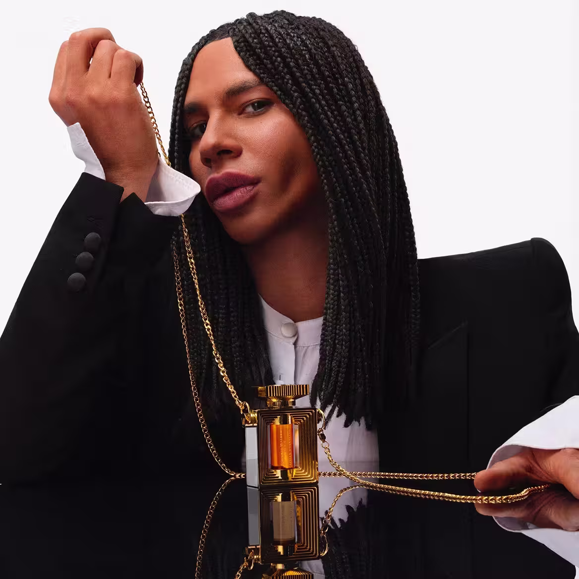 A person with long braided hair, wearing a black blazer and white shirt, holds a gold chain attached to a golden perfume bottle. The bottle rests on a reflective black surface against a plain white background.
