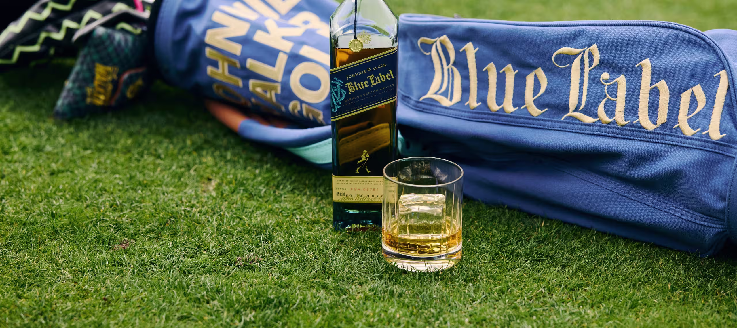 A bottle of whisky and a glass with a golf ball-shaped ice cube sit on a golf courses green grass. Trees and sand bunkers are visible in the blurred background.