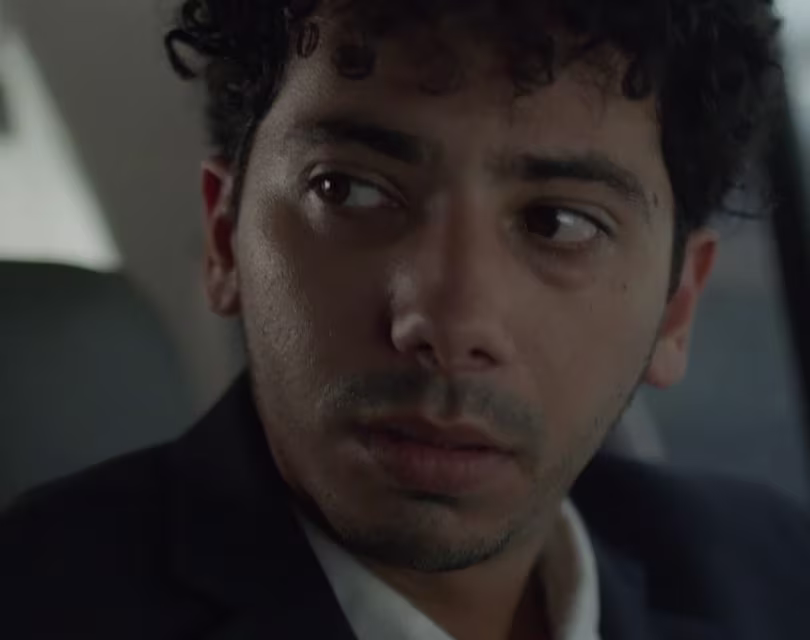 Una persona de pelo rizado y barba fina está sentada en un coche, vistiendo una chaqueta oscura y mirando pensativa hacia un lado. La imagen transmite una expresión pensativa o preocupada.